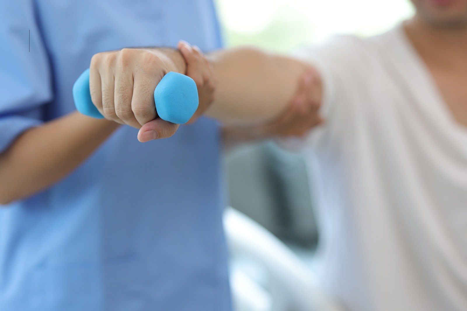 Nurse in an examination room at a hospital is doing physiotherapy for a male patient's arm with dumbbells lifting up and down.