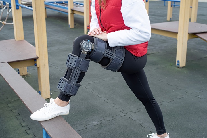 Woman wearing sport clothes and knee brace or orthosis after leg surgery, walking down the stairs in the park. Medical and healthcare concept.