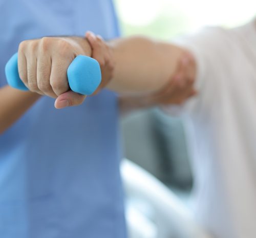Nurse in an examination room at a hospital is doing physiotherapy for a male patient's arm with dumbbells lifting up and down.